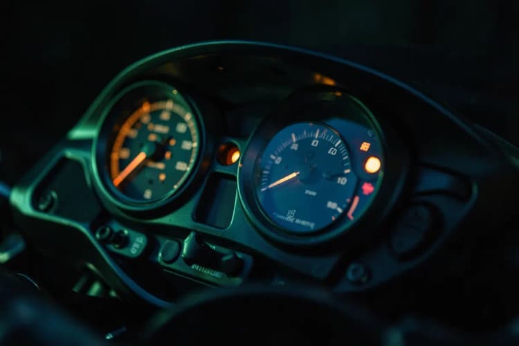 Close-up de um painel de motocicleta com velocímetro e luzes indicadoras apagadas, em um ambiente escuro.